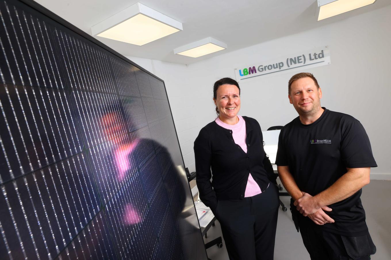 wo people stand indoors next to a large solar panel, smiling at the camera. A sign behind them reads LBM Group (NE) Ltd.