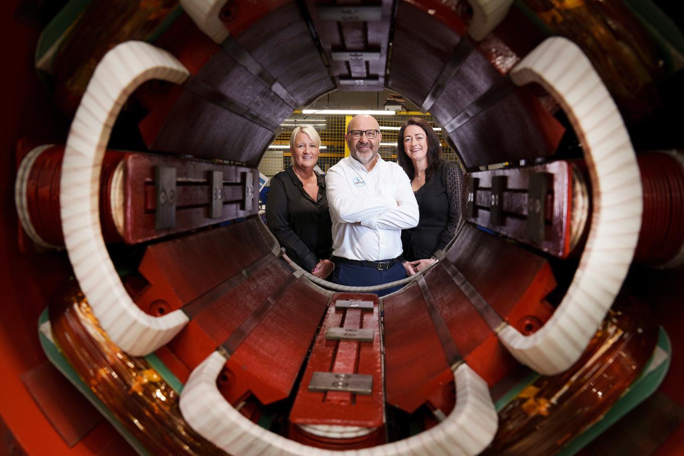 Three people stand inside a large industrial machine, smiling at the camera